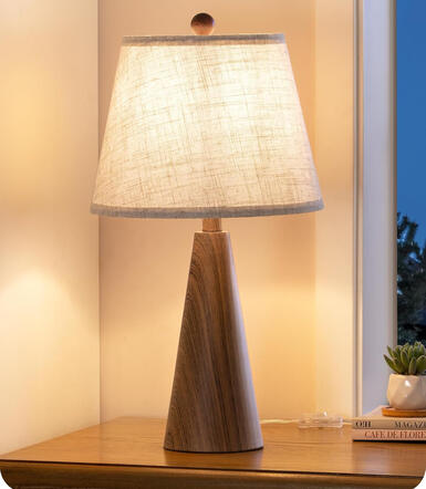 Boho-inspired table lamp with warm beige linen shade and faux woodgrain metal base. A timeless accent for cozy spaces. Small beige linen table lamp with woodgrain-look metal base on light wood nightstand beside cozy bed.