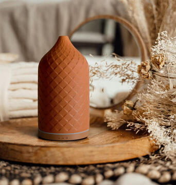 Ground your space with calming scent and sculptural design — a functional accent that complements any cozy bedroom. Warm and cozy essential oil diffuser emitting soft mist on a nightstand, styled beside a candle and book.