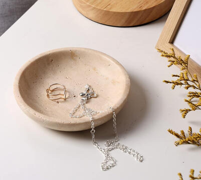 Small round travertine tray holding rings and a gold bracelet on a wood bedside table with soft morning light.