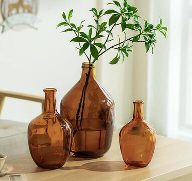 Amber recycled glass vase with curved shape and narrow neck, displayed with dried stems on a wooden nightstand.