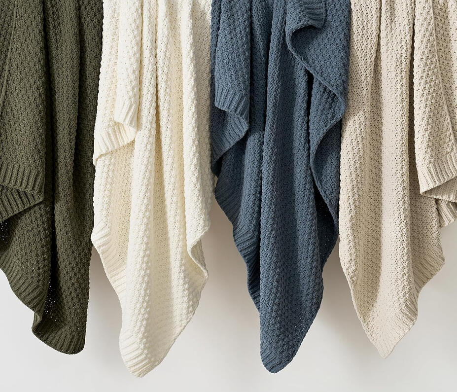 Neutral moss stitch chenille throw blanket folded neatly on a bed with layered pillows in ivory and beige tones.