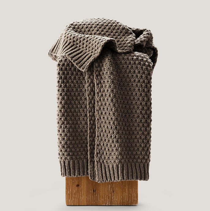 Soft chenille moss-stitch blanket in warm neutral tone, folded at the edge of a bed. Adds cozy texture and subtle mid-century detail.