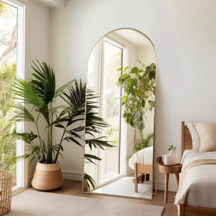 Arched full-length floor mirror with thin metal frame leaning against wall in cozy neutral bedroom or reading nook.