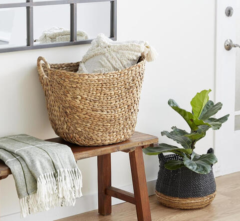 Handwoven from natural seagrass, this roomy basket keeps pillows and blankets close while adding texture and charm. Large woven seagrass basket with arched handles filled with blankets beside reading chair in cozy corner.