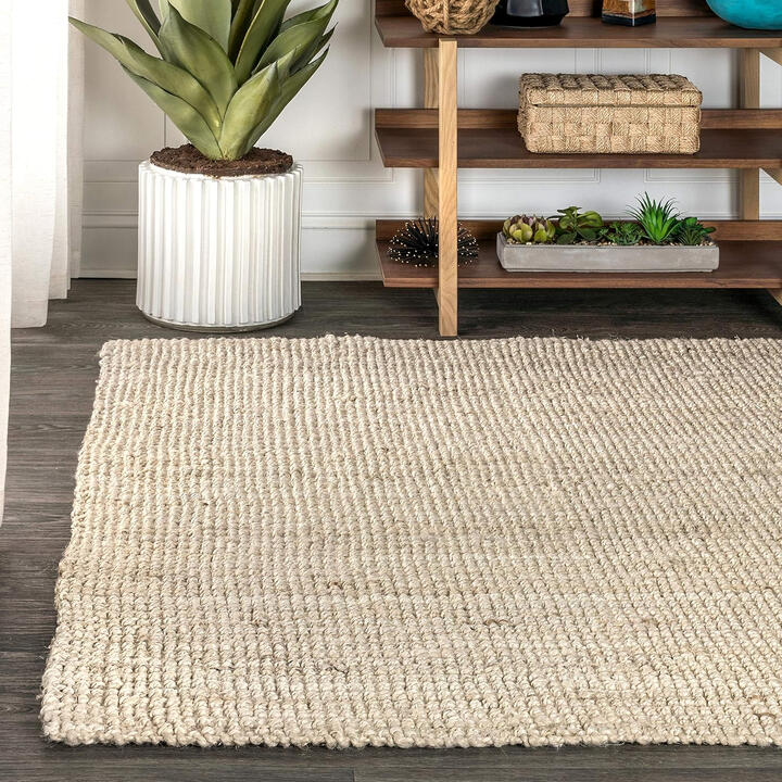 Handwoven natural jute area rug with braided texture in light ivory tone, styled under boho wood bed with neutral bedding.