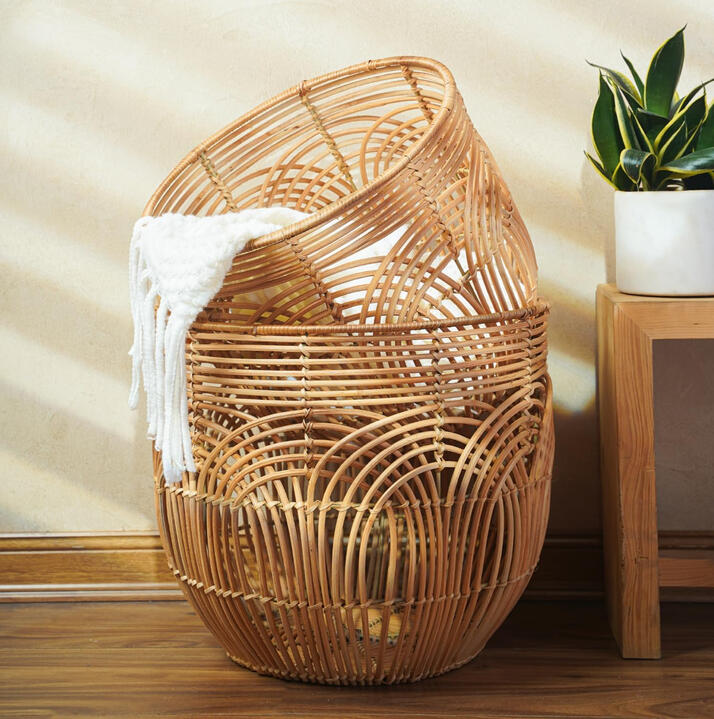 Handwoven natural rattan blanket basket with open weave design, styled beside a bed with neutral textiles. Adds warmth and mid-century organic texture to a bedroom.